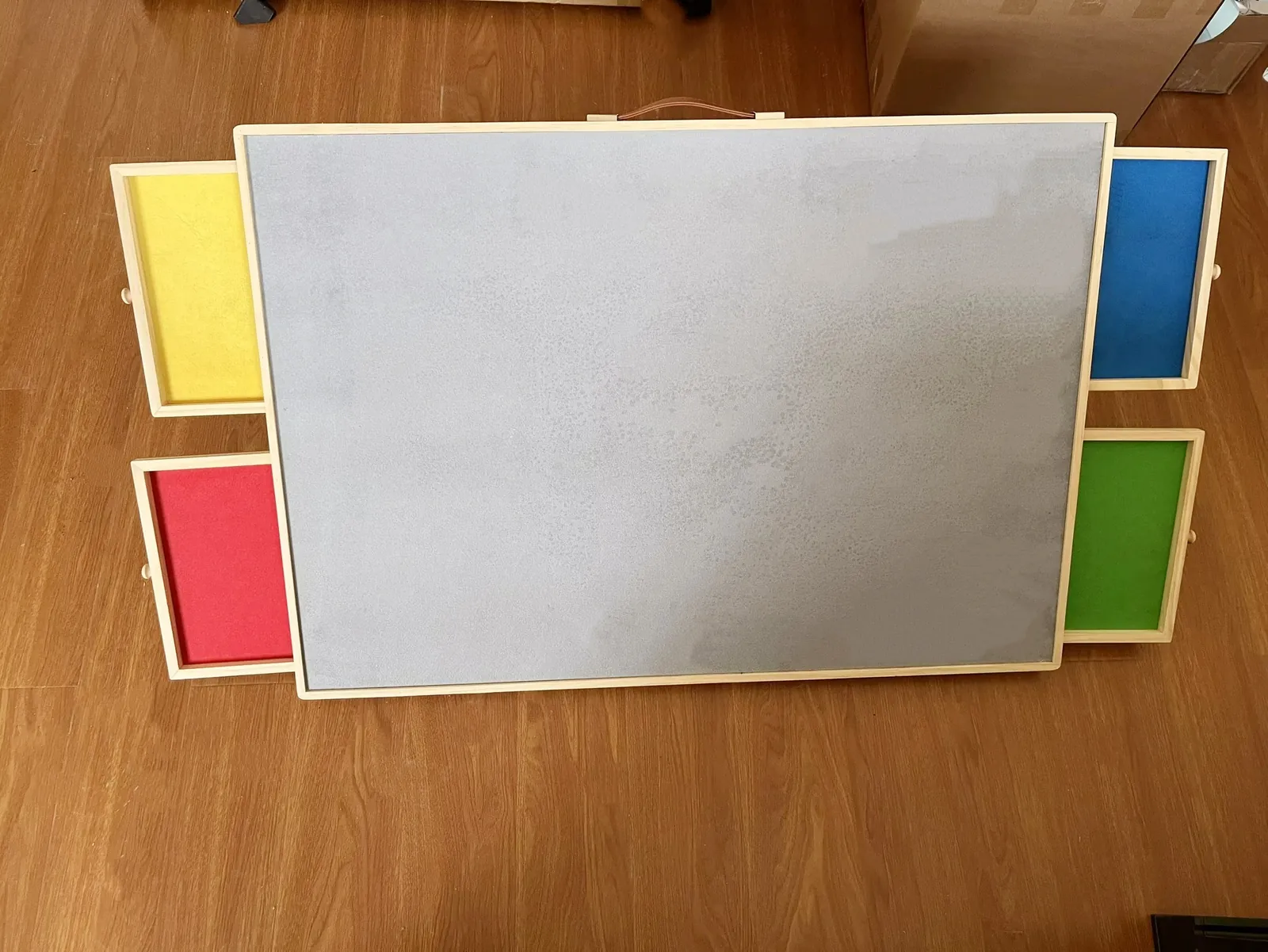 Wooden puzzle board with grey felt surface and four colour-lined drawers on a wood floor